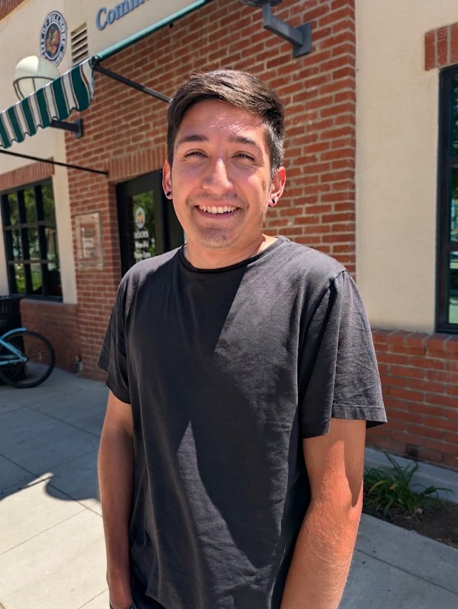 Smiling young man with dark hair and black t-shirt stands outside a red brick building.