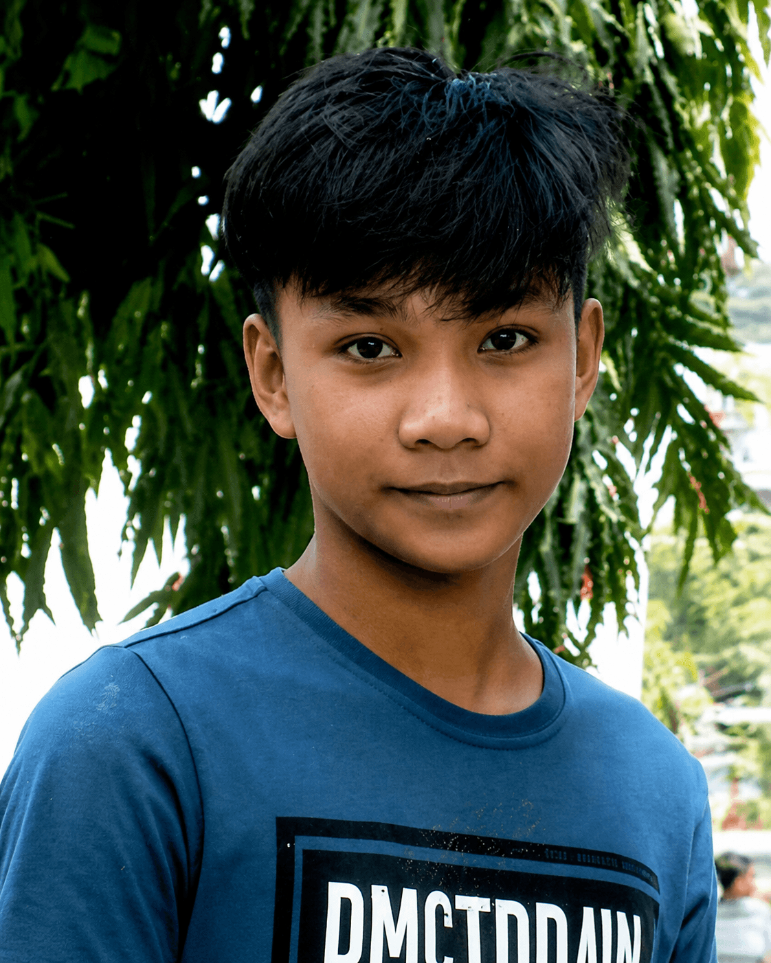 Young man with dark hair and a blue shirt posing against lush green foliage.